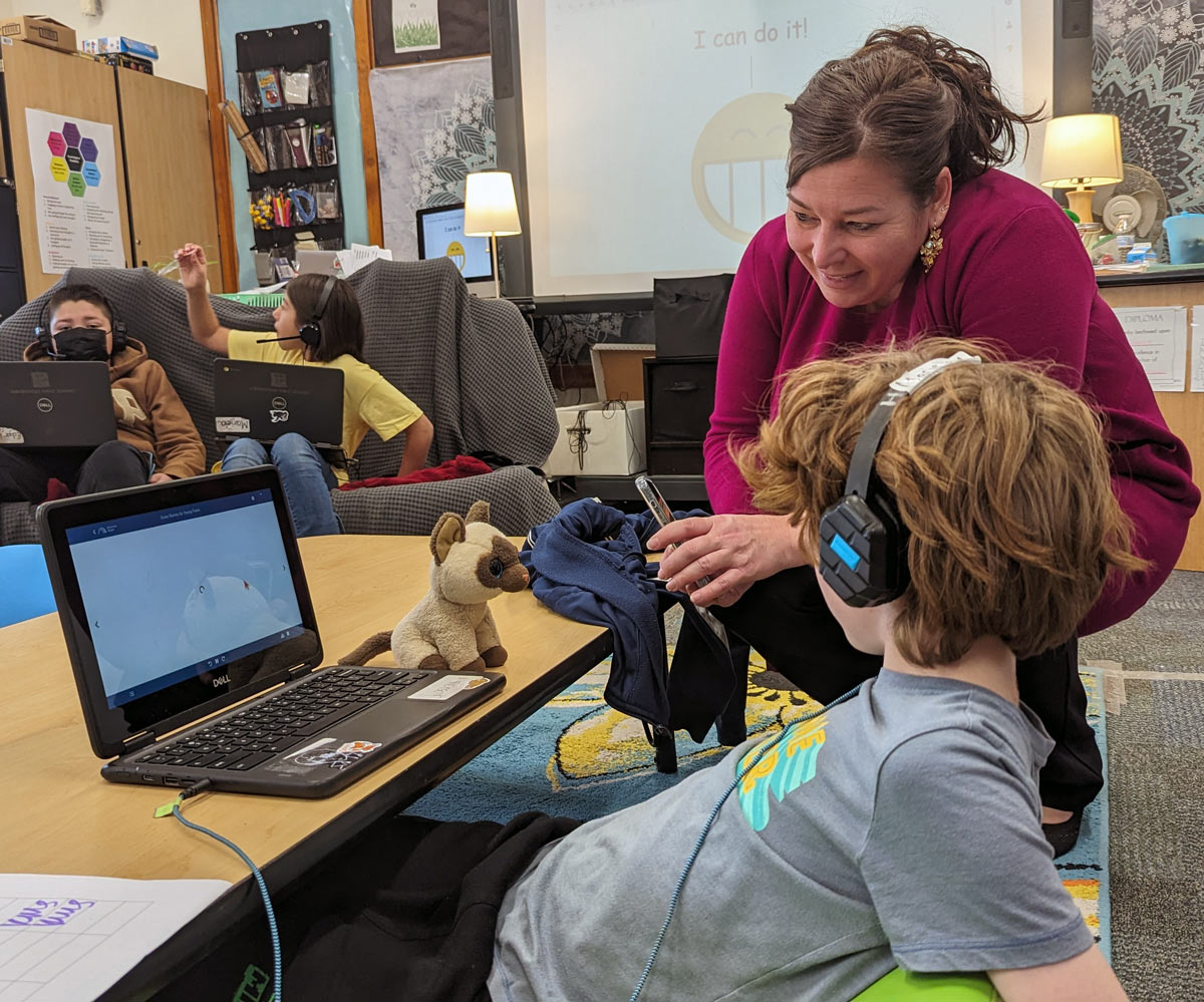 Photo of teacher next to young boy on computer with headphones
